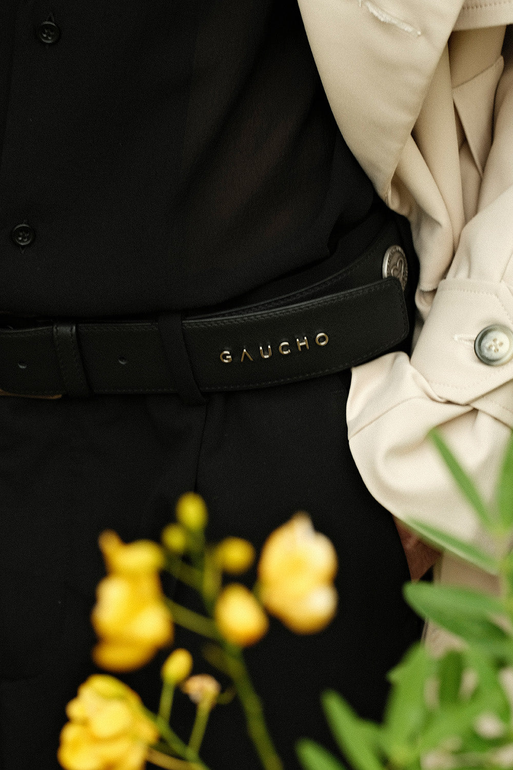 Close-up of a person wearing a black belt with 'Gaucho' branding, surrounded by yellow flowers and a beige garment.