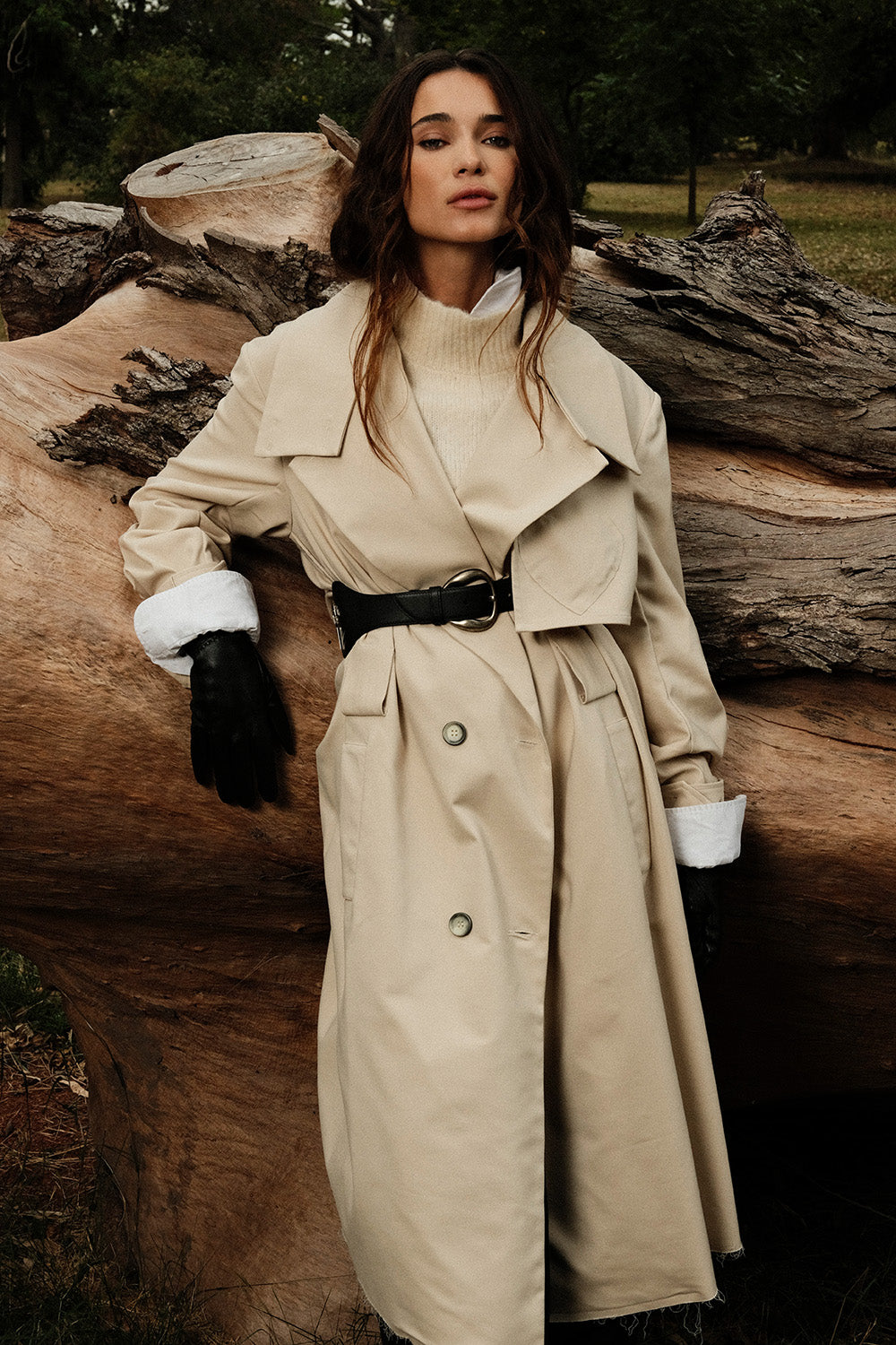 Woman in a beige trench coat standing among large logs in a forest setting