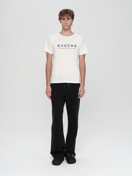 Person wearing a white t-shirt with 'Gaucho' logo and black pants on a white background