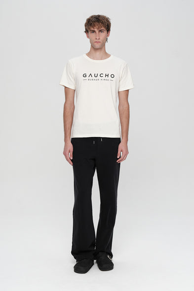 Person wearing a white t-shirt with 'Gaucho' logo and black pants on a white background