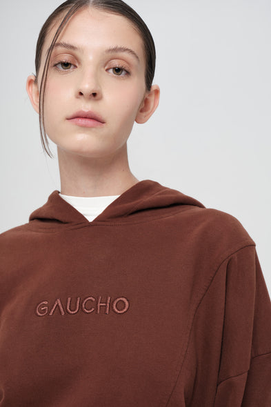 Person wearing a brown hoodie with 'GAUCHO' branding on a plain background