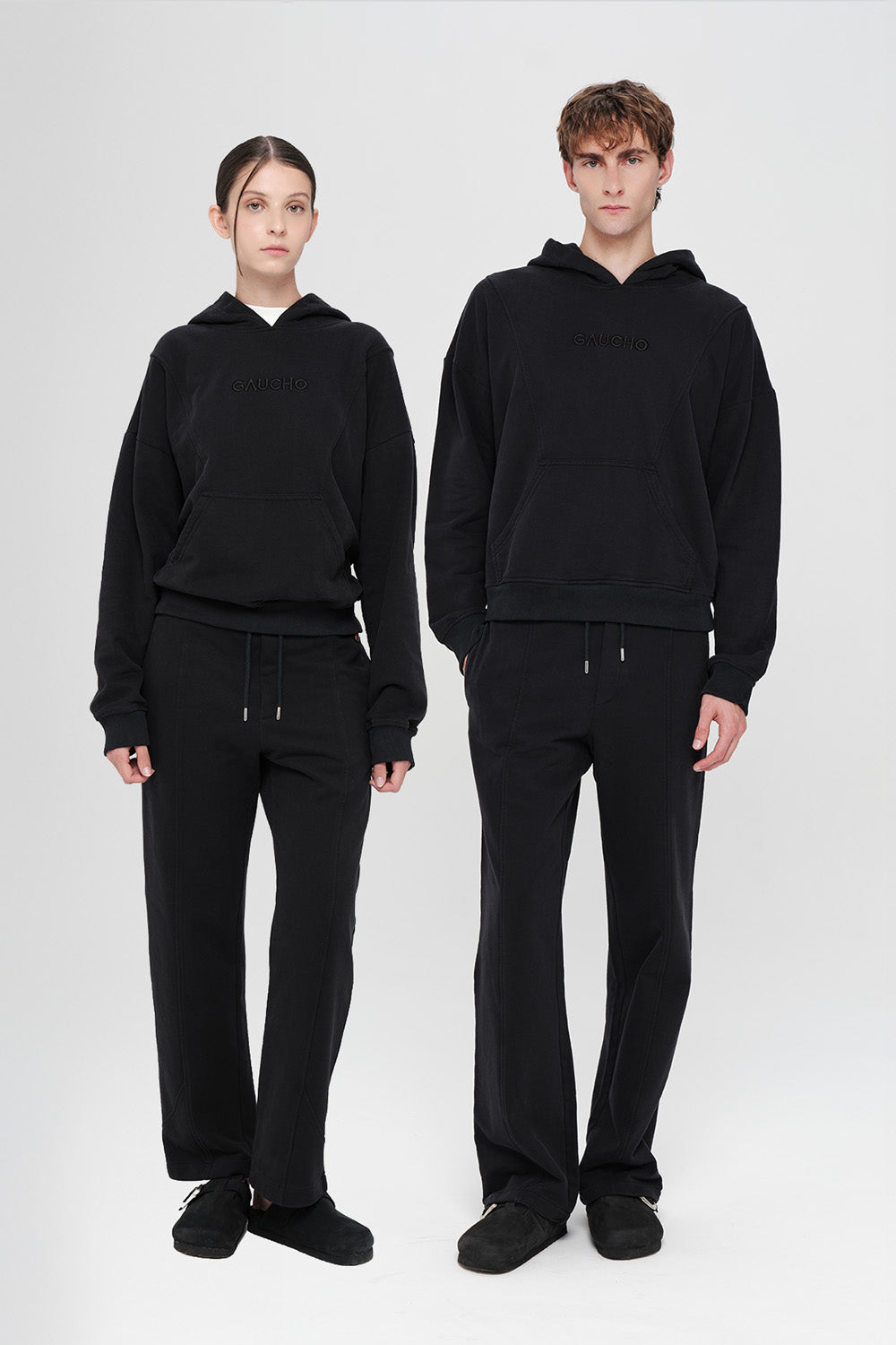 Two people wearing matching black hoodies and pants on a white background
